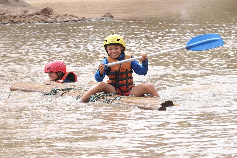 Two children wearing helmets and life jackets enjoy rafting on a bamboo raft in a muddy river, smiling and paddling with blue oars.