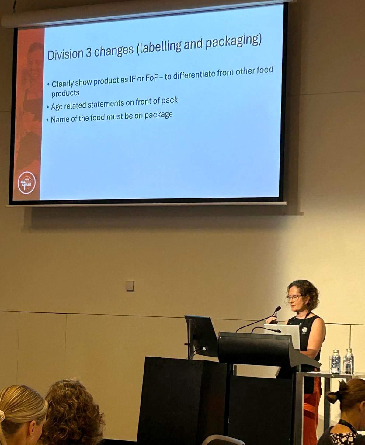 A woman presents at a conference, standing at a podium beneath a slide about Division 3 changes in labeling and packaging.