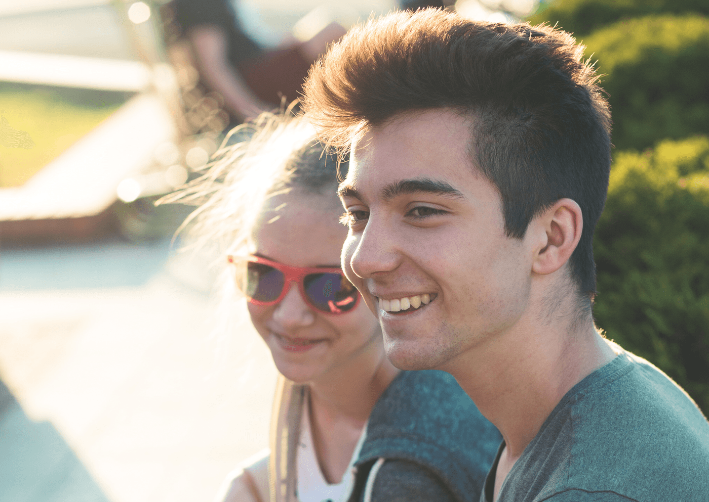 A young man and woman sitting outdoors, smiling. The woman wears red sunglasses, and sunlight highlights their faces.