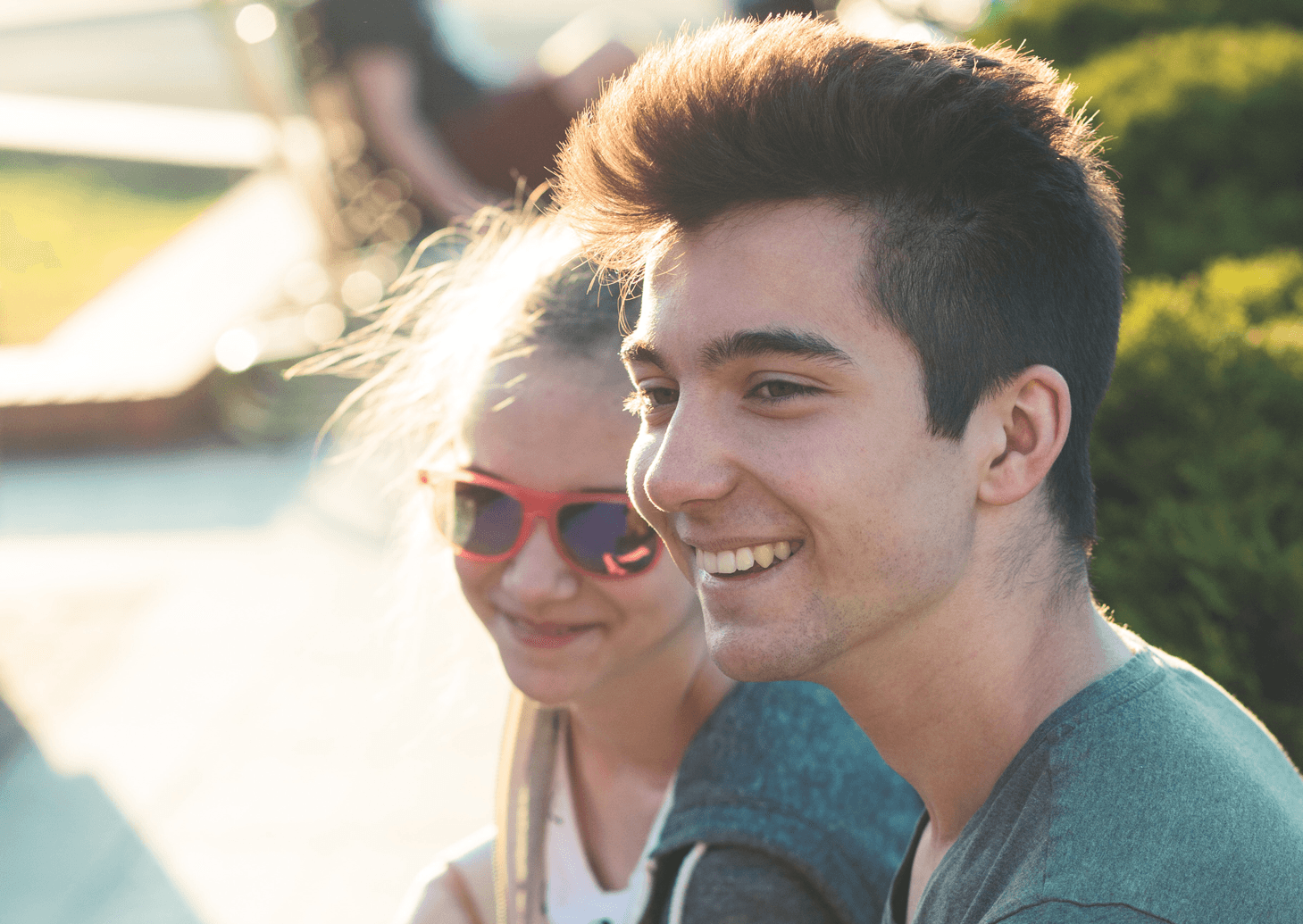 A young man and woman sitting outdoors, smiling. The woman wears red sunglasses, and sunlight highlights their faces.