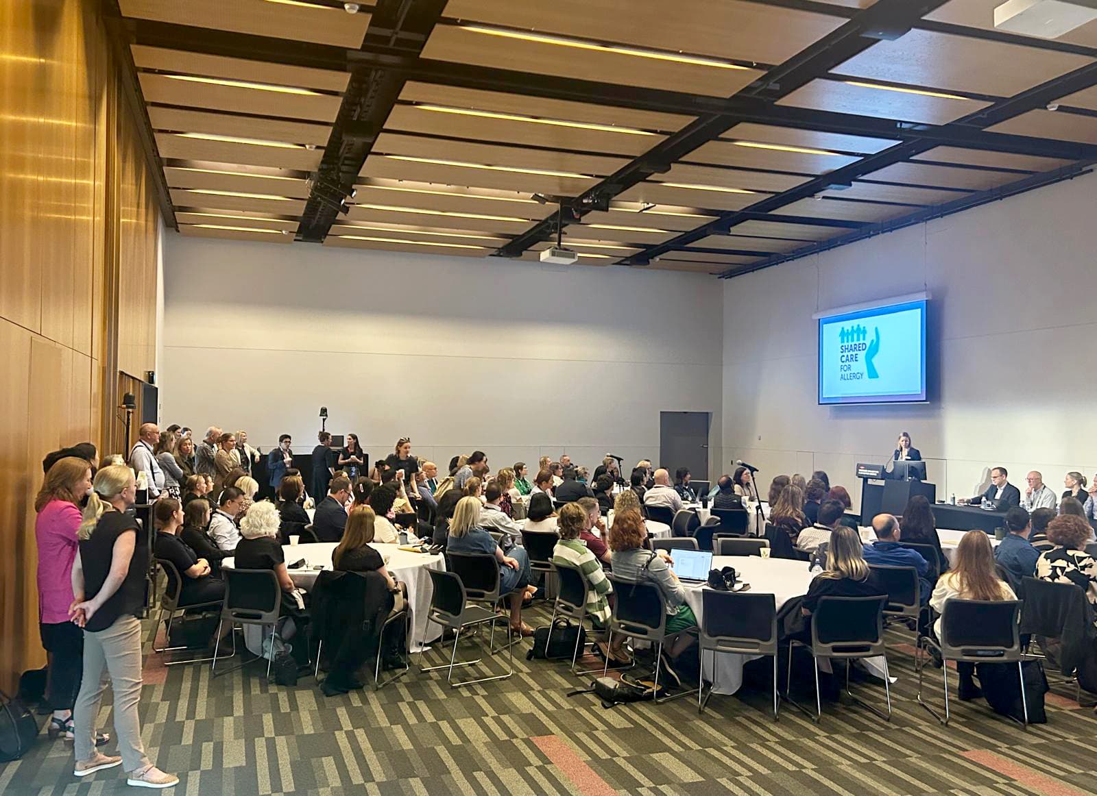 A conference room filled with seated and standing attendees facing a speaker at a podium. A projected presentation is visible on the wall.
