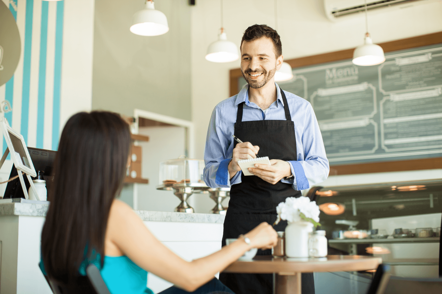A smiling waiter in an apron takes an order from a seated woman in a bright cafe with a menu board and cake display in the background.
