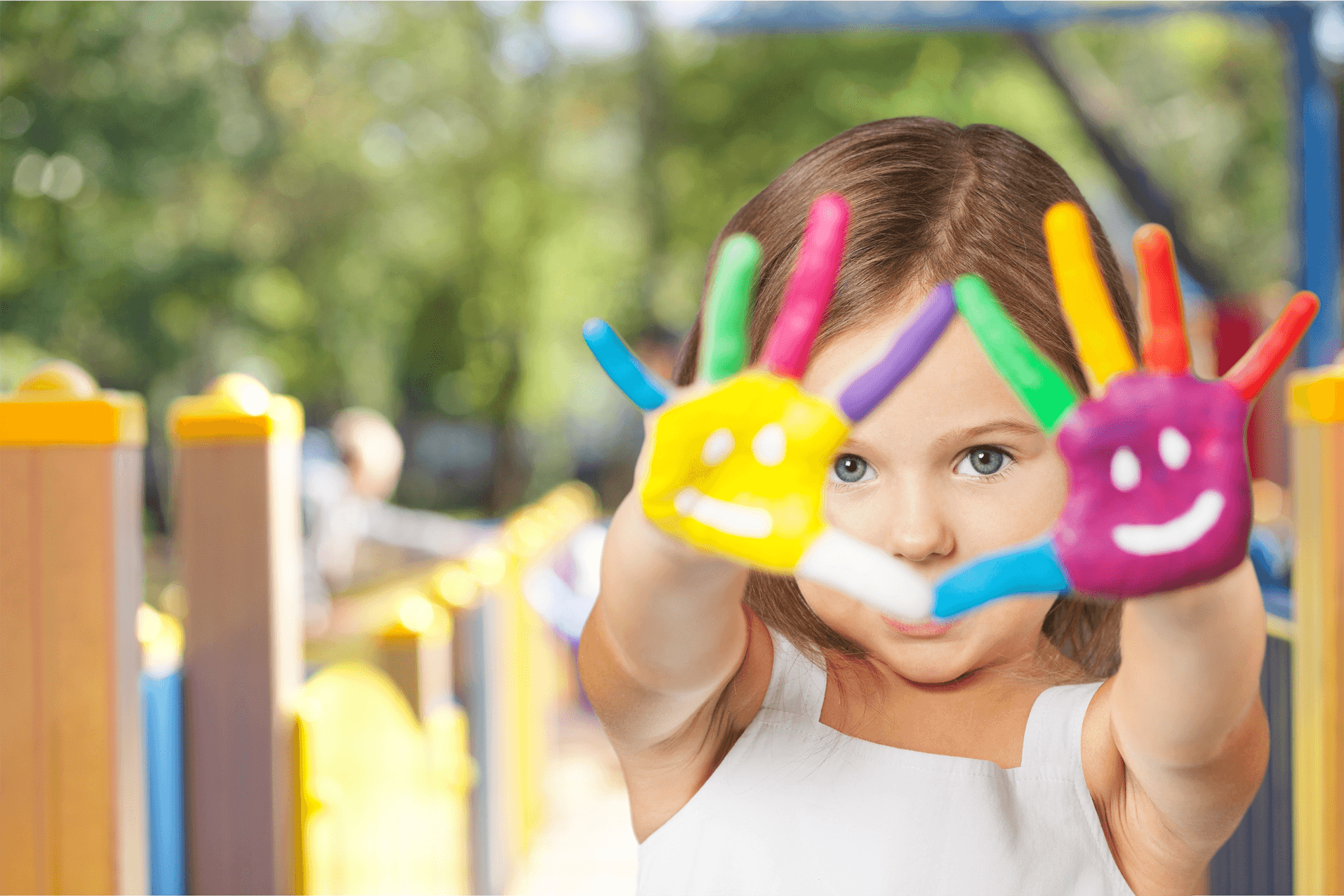 A young girl in a white dress holds up her hands painted with colorful smiley faces, standing in a vibrant playground.