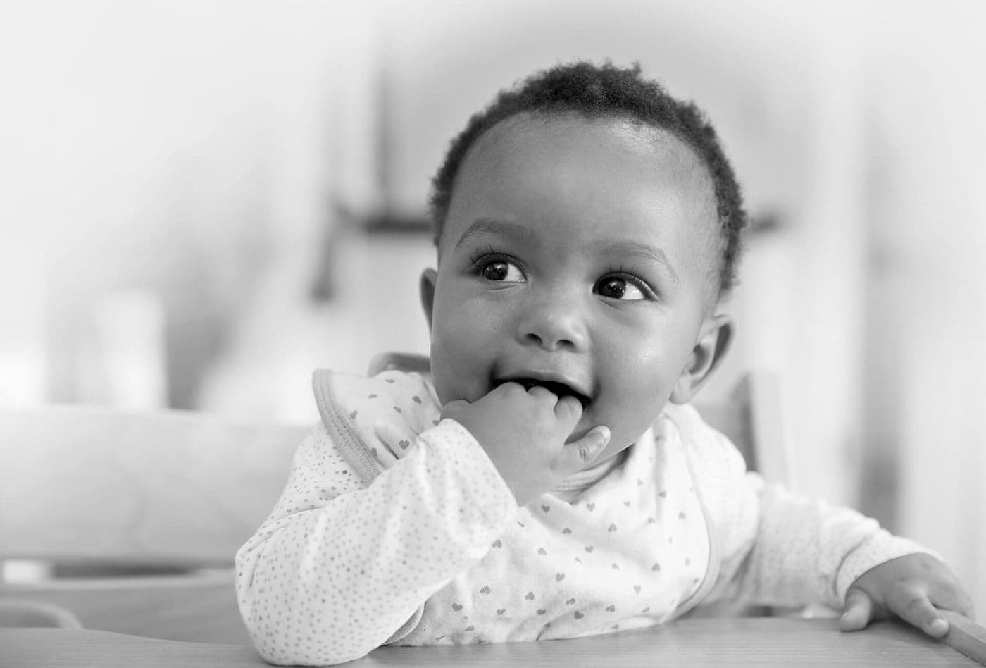Black and white photo of a baby sitting at a table, wearing a bib, with fingers in their mouth and looking to the side.