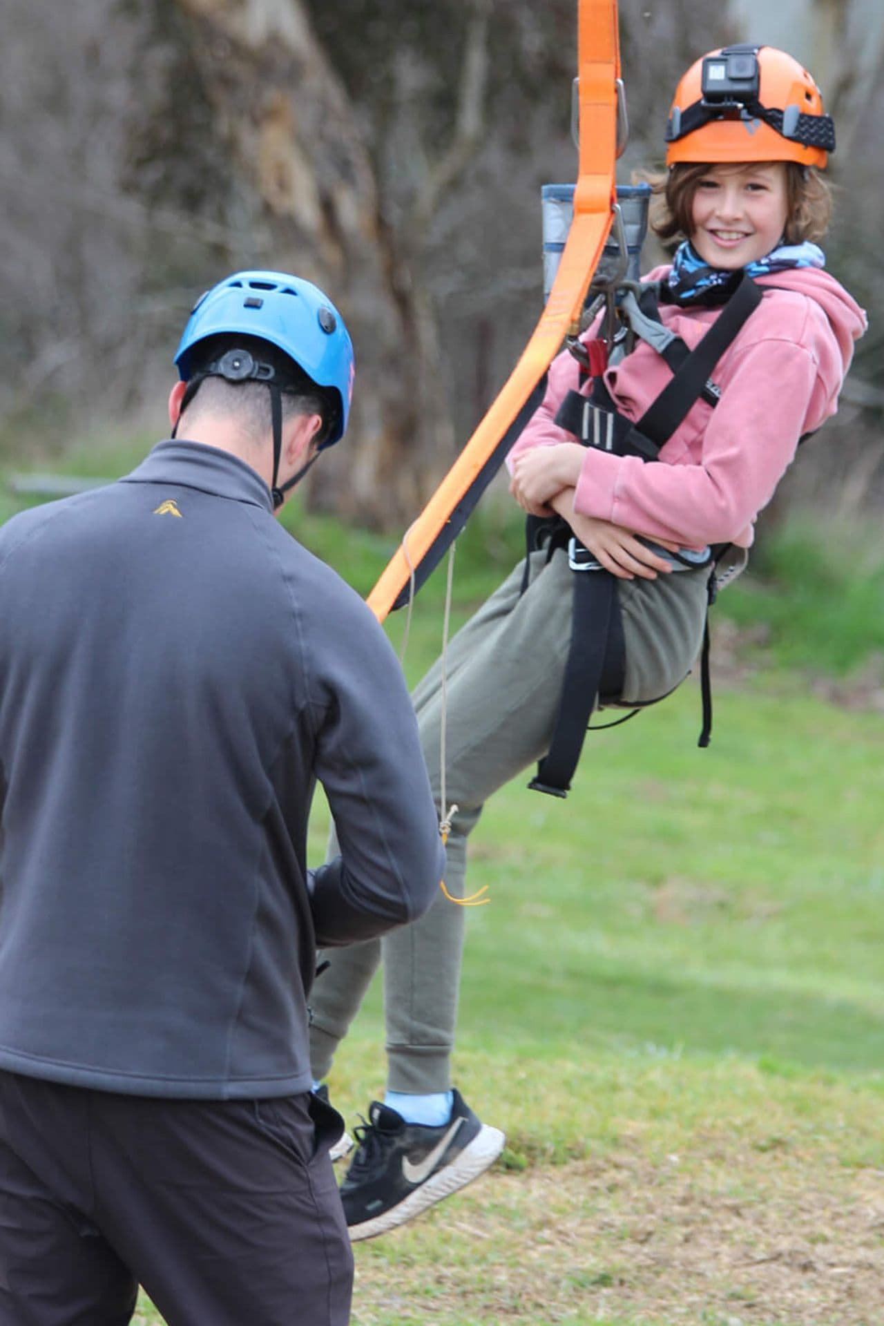 A person in a pink hoodie and helmet is zip-lining, assisted by another person wearing a blue helmet