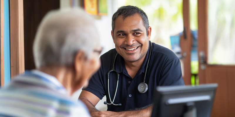 A doctor with a stethoscope smiles while talking to an elderly patient in a brightly lit room.