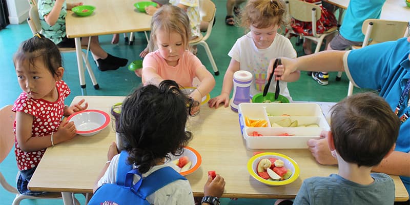 Children sitting around a table in a classroom, eating fruit from bowls. An adult assists with serving from a container.