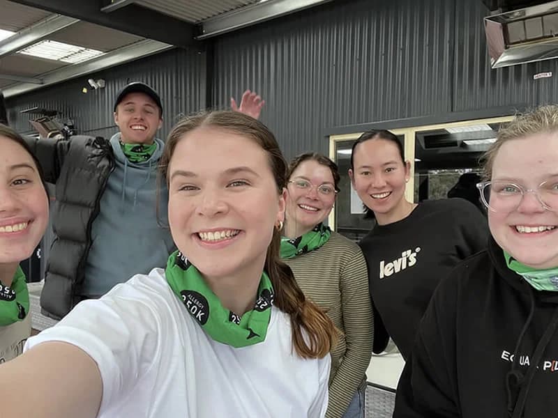 A group of six people smiling indoors, wearing green bandanas. They are casually dressed, with a dark wall and windows in the background.