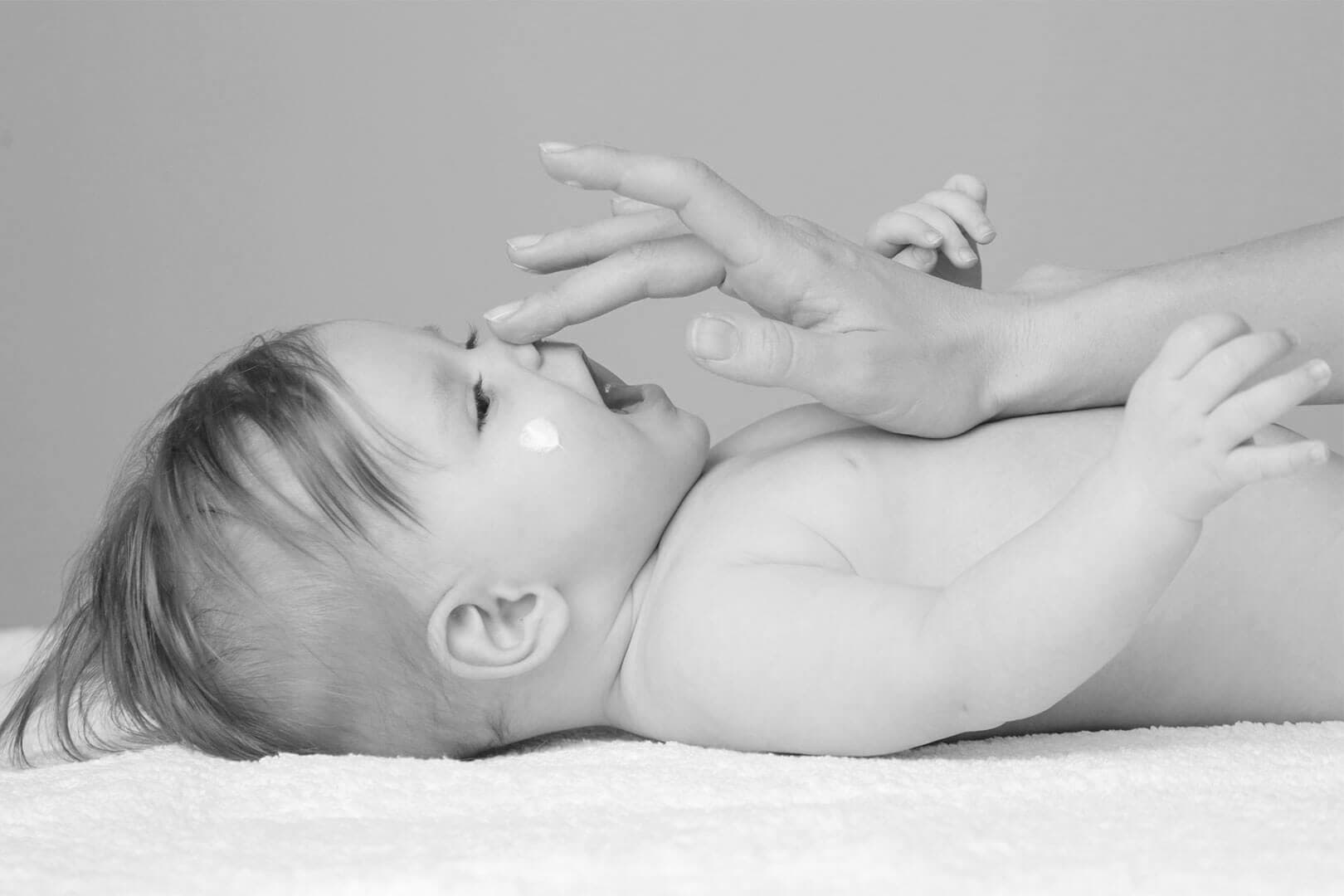 A baby lying on a soft surface smiles as an adult's hand gently touches their nose.