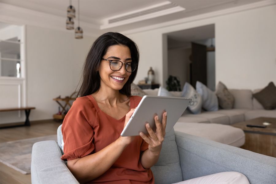 Smiling woman with glasses uses a tablet while sitting on a gray couch in a modern living room.