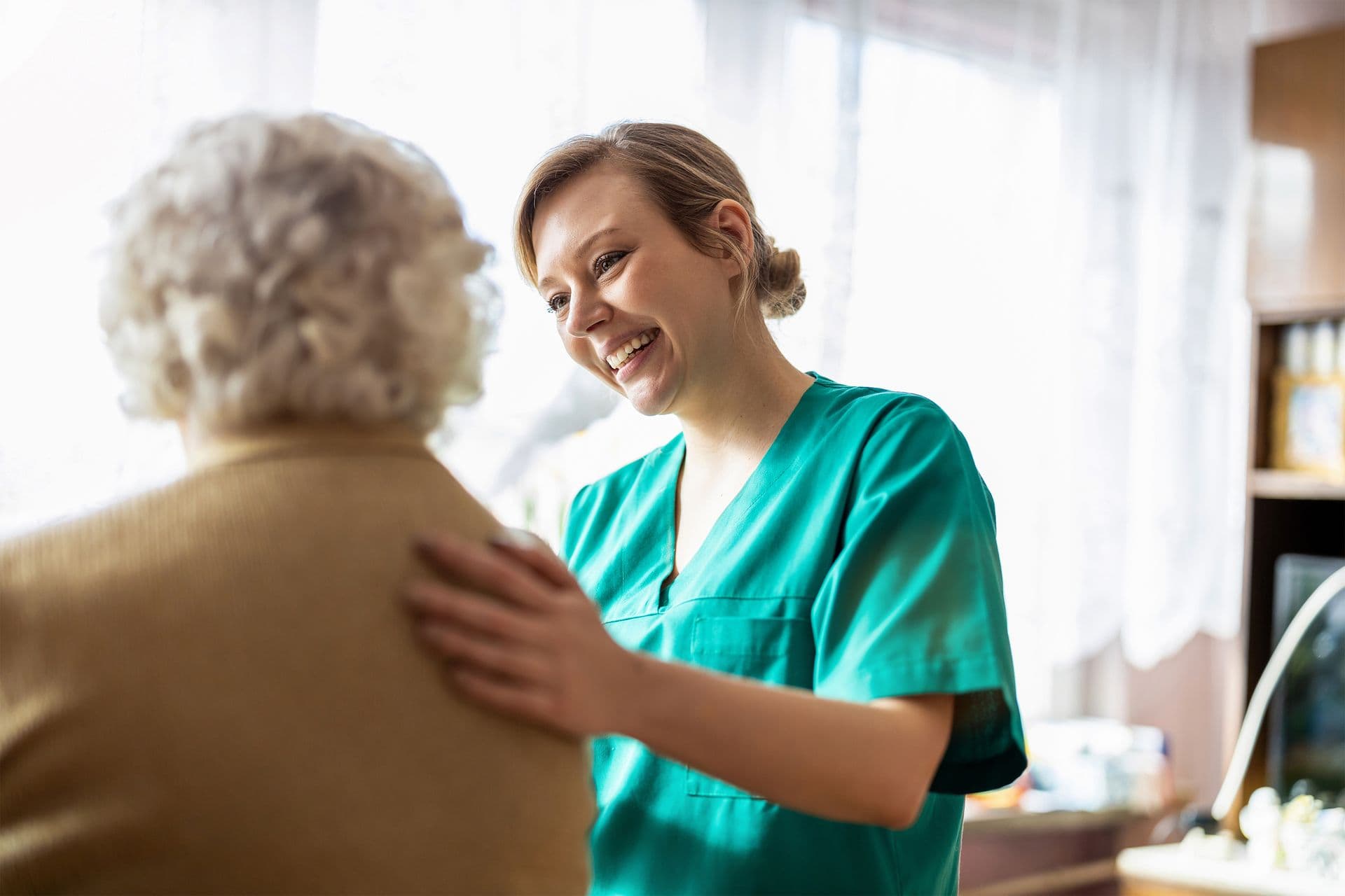 A smiling caregiver in green scrubs gently places a hand on the shoulder of an elderly person, offering comfort in a warmly lit room.