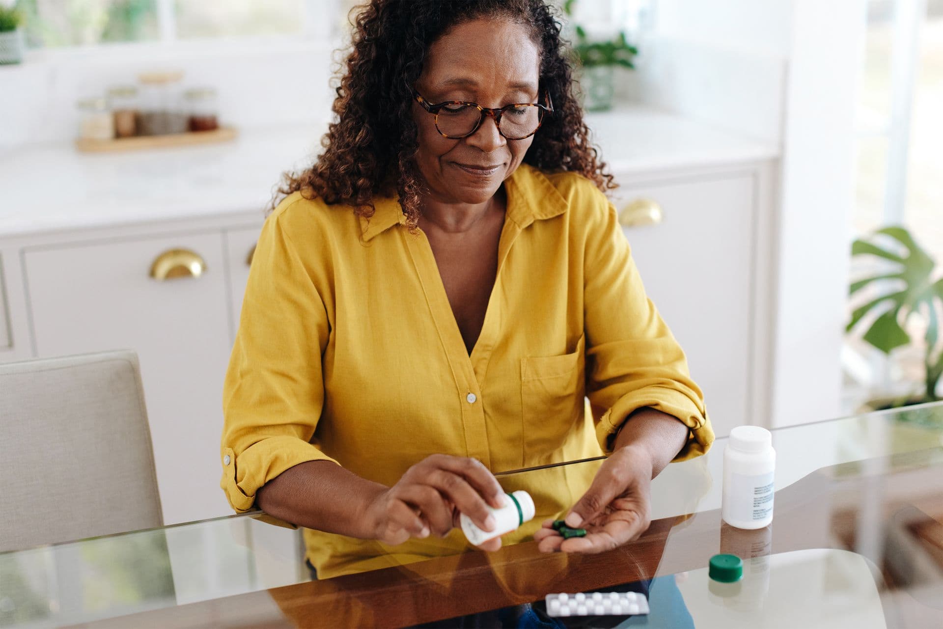 A woman in a yellow shirt sits at a table, dispensing pills from a bottle into her hand, with medication bottles nearby.
