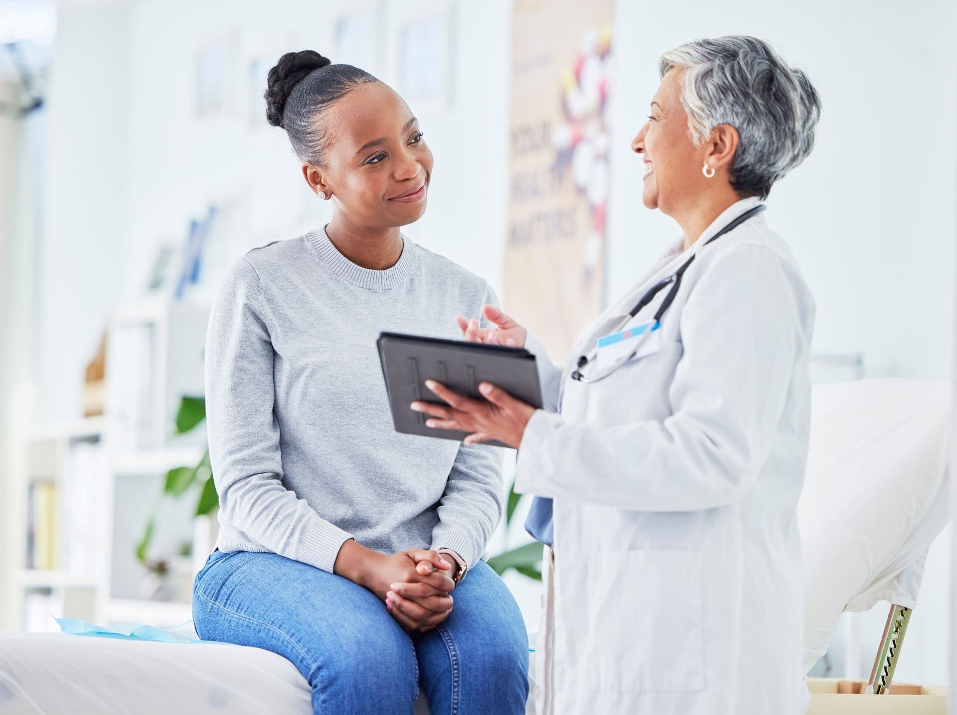 Doctor and patient discussing results in a bright medical office. The patient sits on an examination table, while the doctor holds a tablet.