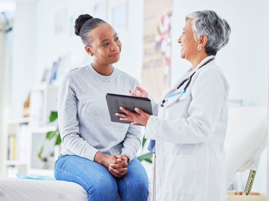 Doctor and patient discussing results in a bright medical office. The patient sits on an examination table, while the doctor holds a tablet.
