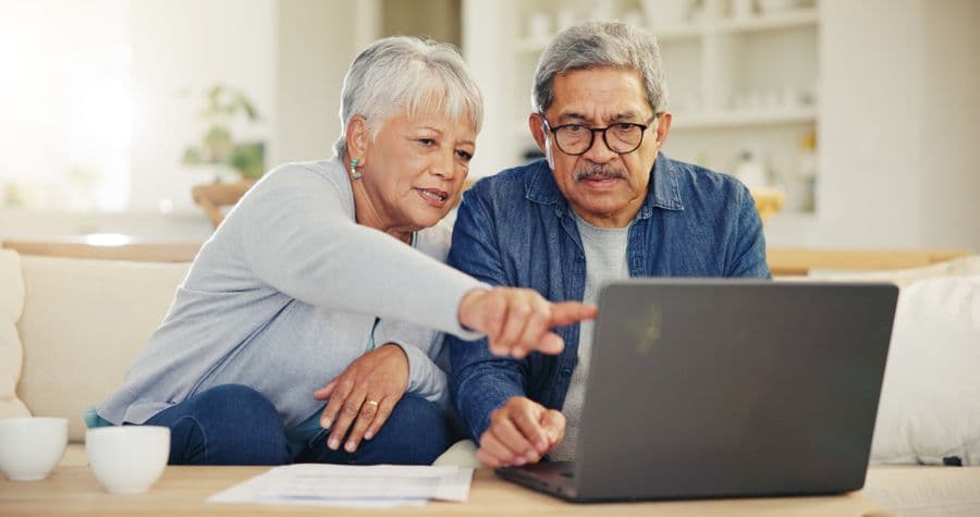 Elderly couple sitting on a couch, looking at a laptop screen. The woman is pointing at the screen, and both appear focused and engaged.