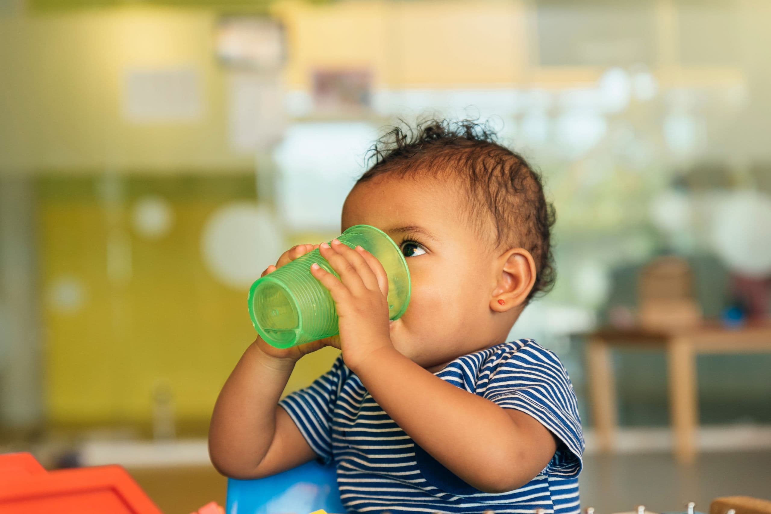 A toddler in a striped shirt drinks from a green cup, sitting indoors.