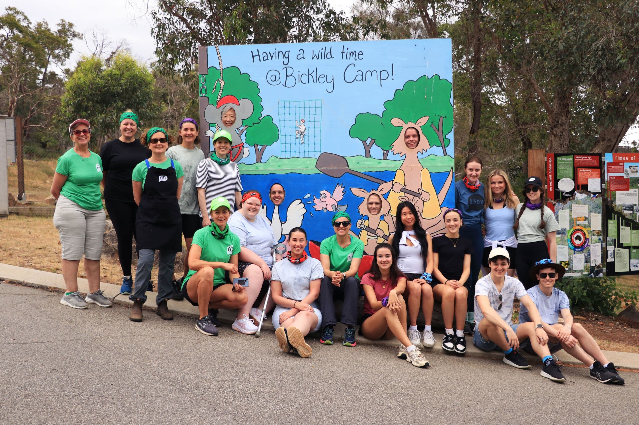 Large group of smiling camp participants posing in front of a colorful "Having a wild time @Bickley Camp!" photo cutout outdoors.