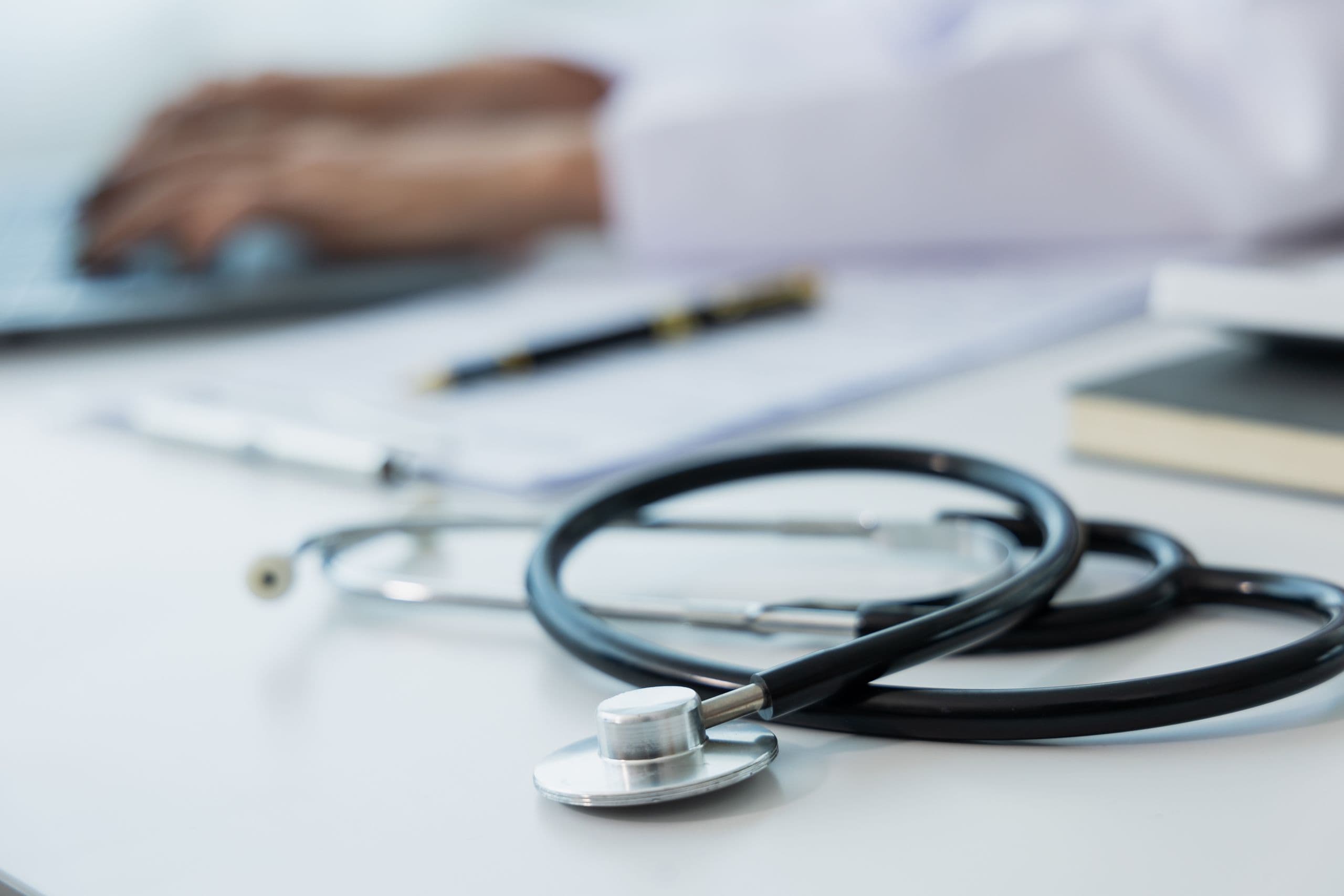Stethoscope on a desk with a laptop keyboard in use by blurred hands, a pen and notebook in the background.