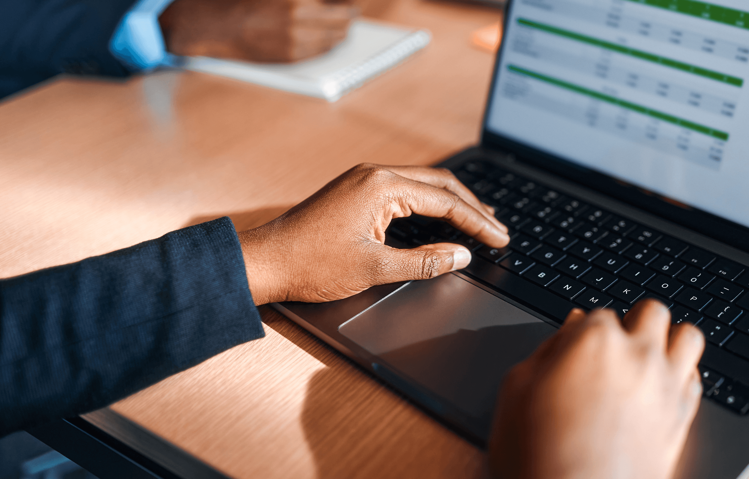 Person typing on a laptop displaying a spreadsheet, with another person taking notes nearby on a wooden desk.