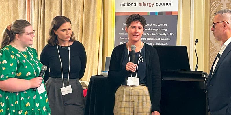 Four people are standing and speaking at an event. One person holds a microphone. A banner reads "national allergy council."